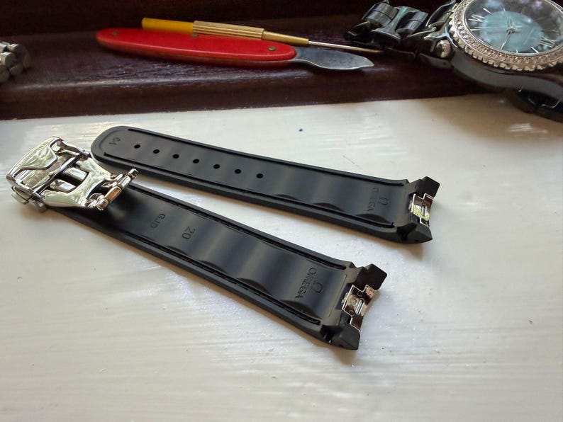 Puede incluir: Una correa de reloj de caucho negro con un cierre desplegable de metal plateado. La correa tiene grabado el nombre de la marca Omega y el n&uacute;mero 20. La banda de dos piezas presenta una superficie texturizada con ranuras y orificios.