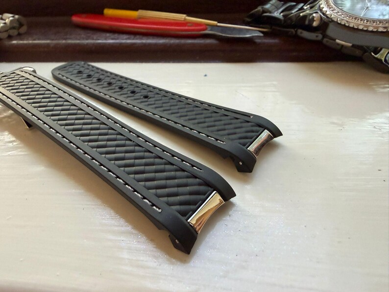 May include: A pair of black rubber watch straps featuring a textured basketweave pattern on the surface, white contrast stitching along the edges, and polished silver-tone metal end links designed for attachment to a watch case.