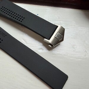May include: A black rubber watch strap featuring a perforated texture and the embossed text TAGHeuer. The strap includes a metallic silver-toned end link with a small, engraved TAGHeuer logo shield, designed for a secure attachment to a watch case.