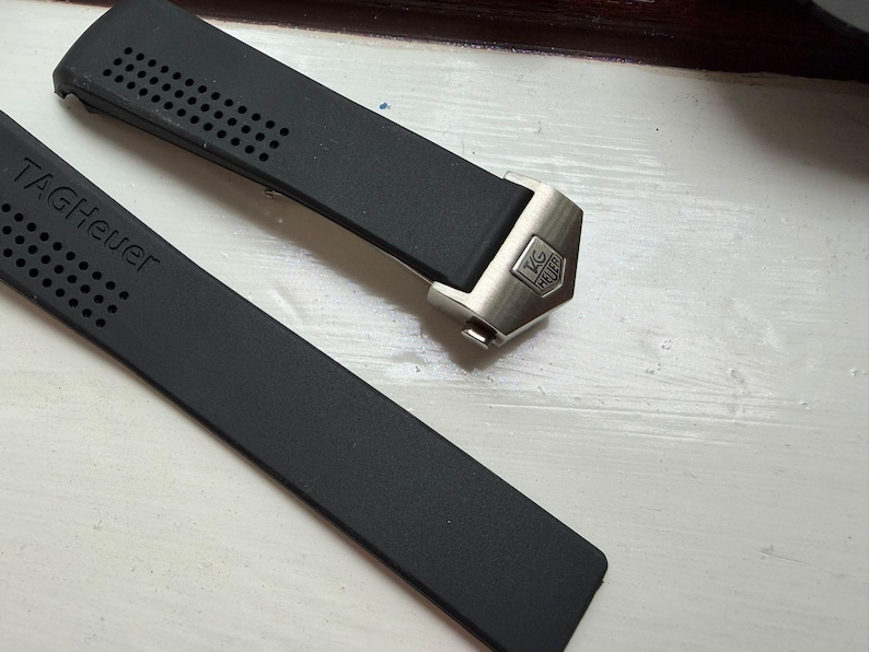 May include: A black rubber watch strap featuring a perforated texture and the embossed text TAGHeuer. The strap includes a metallic silver-toned end link with a small, engraved TAGHeuer logo shield, designed for a secure attachment to a watch case.