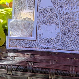 May include: A white laser-cut greeting card featuring a champagne bottle and glass silhouette with intricate damask patterns. The card is embellished with a white lace heart, silver-tipped pins, and a row of small pink pearls along the bottom edge.