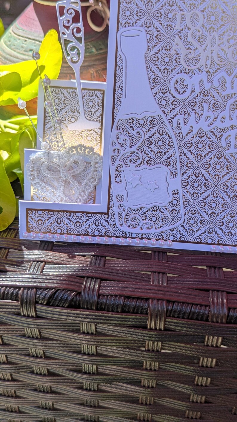 May include: A white laser-cut greeting card featuring a champagne bottle and glass silhouette with intricate damask patterns. The card is embellished with a white lace heart, silver-tipped pins, and a row of small pink pearls along the bottom edge.