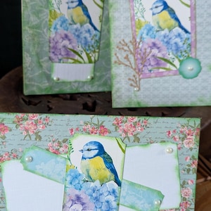 May include: Three handmade greeting cards featuring blue and yellow birds perched on purple and blue hydrangea flowers. The cards are decorated with floral patterned paper, script text, and small white pearl accents on layered paper tags.