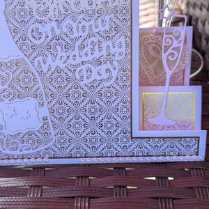 May include: A wedding greeting card featuring a gold and white intricate damask pattern, die-cut champagne flute accents, and white pearl embellishments. The text reads To a Special Couple on your Wedding Day in elegant white script lettering.