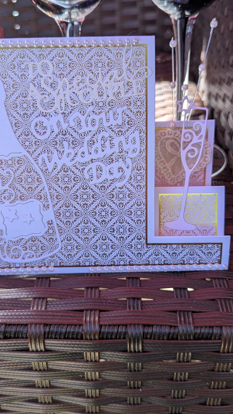 May include: A wedding greeting card featuring a gold and white intricate damask pattern, die-cut champagne flute accents, and white pearl embellishments. The text reads To a Special Couple on your Wedding Day in elegant white script lettering.