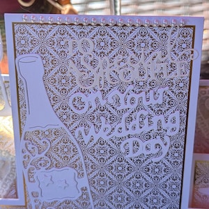 May include: A square wedding greeting card featuring a white and gold damask pattern background. The card displays a die-cut white champagne bottle with star accents and the text To a Special Couple on Your Wedding Day. Small white pearls line the top and bottom edges of the card.