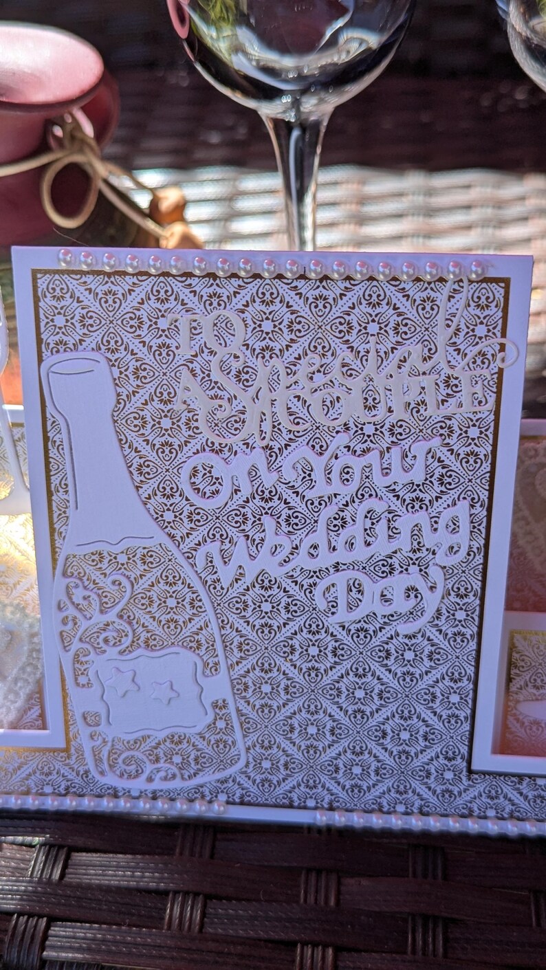 May include: A square wedding greeting card featuring a white and gold damask pattern background. The card displays a die-cut white champagne bottle with star accents and the text To a Special Couple on Your Wedding Day. Small white pearls line the top and bottom edges of the card.