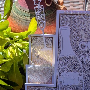 May include: A white and gold greeting card featuring a die-cut champagne bottle and a flute glass. The card has an intricate damask pattern, a white lace heart embellishment, and small pearl accents along the edges, creating a layered design.