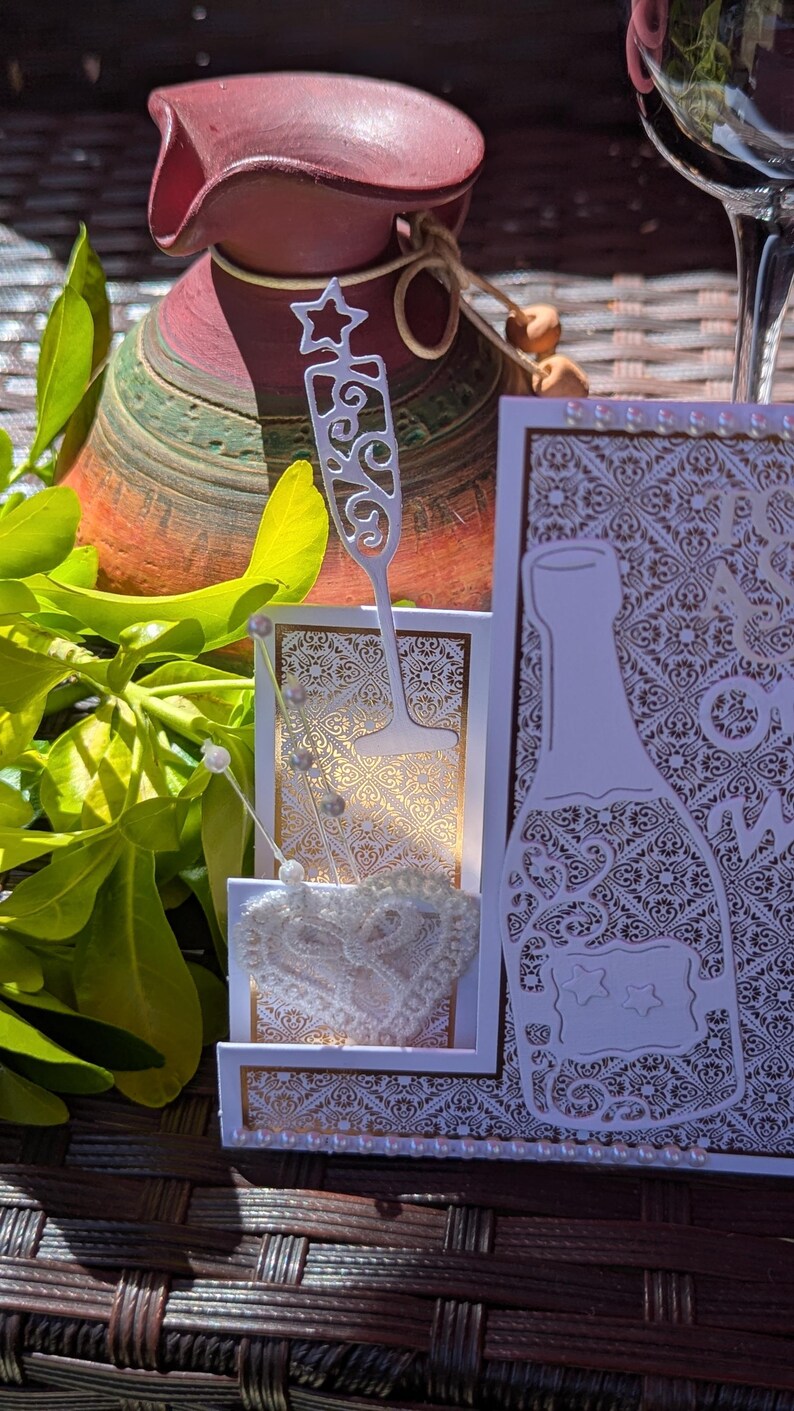 May include: A white and gold greeting card featuring a die-cut champagne bottle and a flute glass. The card has an intricate damask pattern, a white lace heart embellishment, and small pearl accents along the edges, creating a layered design.