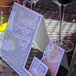 May include: A white wedding greeting card featuring intricate laser-cut patterns, a champagne bottle silhouette, and the text On Your Wedding Day. The card is accented with small white pearls and a delicate lace heart detail on the front panel.