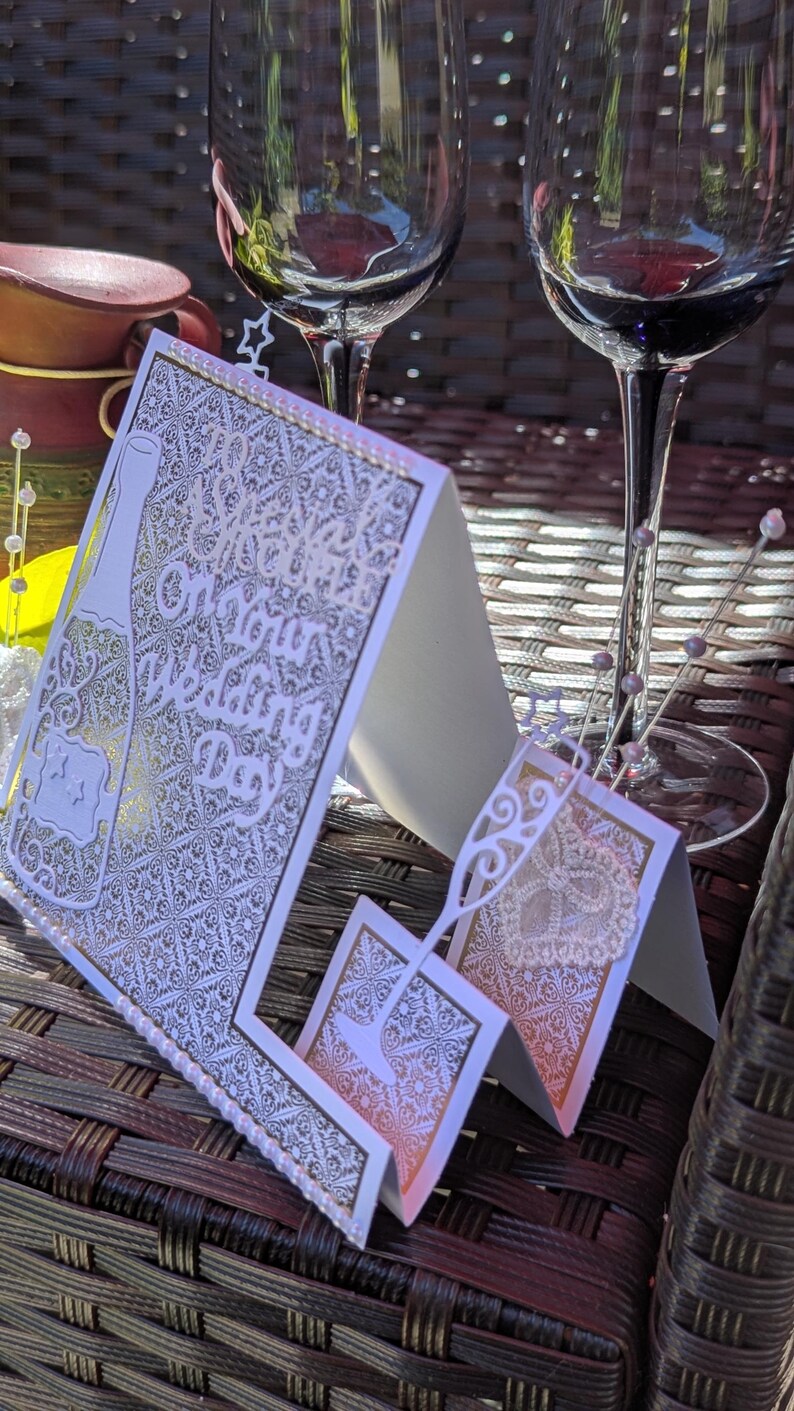 May include: A white wedding greeting card featuring intricate laser-cut patterns, a champagne bottle silhouette, and the text On Your Wedding Day. The card is accented with small white pearls and a delicate lace heart detail on the front panel.