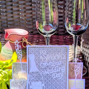 May include: A decorative wedding card featuring a white die-cut champagne bottle and two champagne flutes with the text To Sparkle On Your Wedding Day. The card has a gold-bordered, white patterned background with small pearl accents.