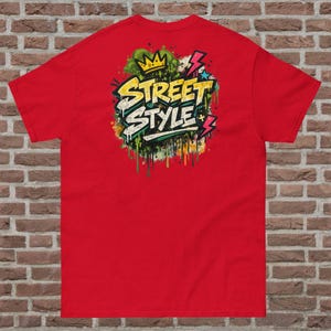 Może przedstawiać: Czerwona koszulka z graficznym napisem "Street Style" w stylu graffiti. Grafika zawiera koronę, błyskawice i krople farby w kolorach zielonym, żółtym i białym. Koszulka wykonana jest z miękkiego materiału.