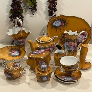 May include: A collection of antique, hand-painted porcelain tea set pieces. The set includes a teapot, creamer, sugar bowl, cups, saucers, a serving platter, and a vase. The pieces are primarily mustard yellow with floral and blue accents.