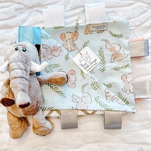Op de afbeelding: Een babyknuffel met een pluche olifant en een vierkant label. Het label is lichtblauw met een olifanten- en bladerenpatroon, en heeft diverse gekleurde linten en een label met de tekst "Three little Snugs Ready for Hugs".
