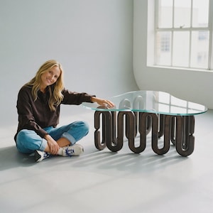 May include: A coffee table featuring a clear, organic-shaped glass top supported by a dark brown wooden base with a continuous, repeating serpentine or wave-like structure that forms the legs and frame of the furniture piece.