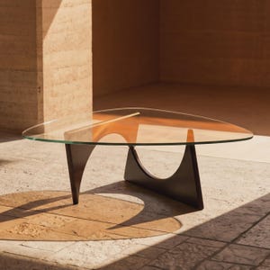 Può includere: Un tavolino da caffè con un piano triangolare in vetro trasparente dai bordi arrotondati. La base è costituita da due supporti in legno curvo di colore marrone scuro che si incrociano per creare una base stabile per la superficie in vetro.