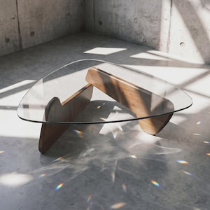 May include: A coffee table featuring a clear, triangular glass top supported by two interlocking, curved wooden base pieces. The wood has a warm, medium-brown tone with visible grain, and the glass top has smooth, rounded edges.