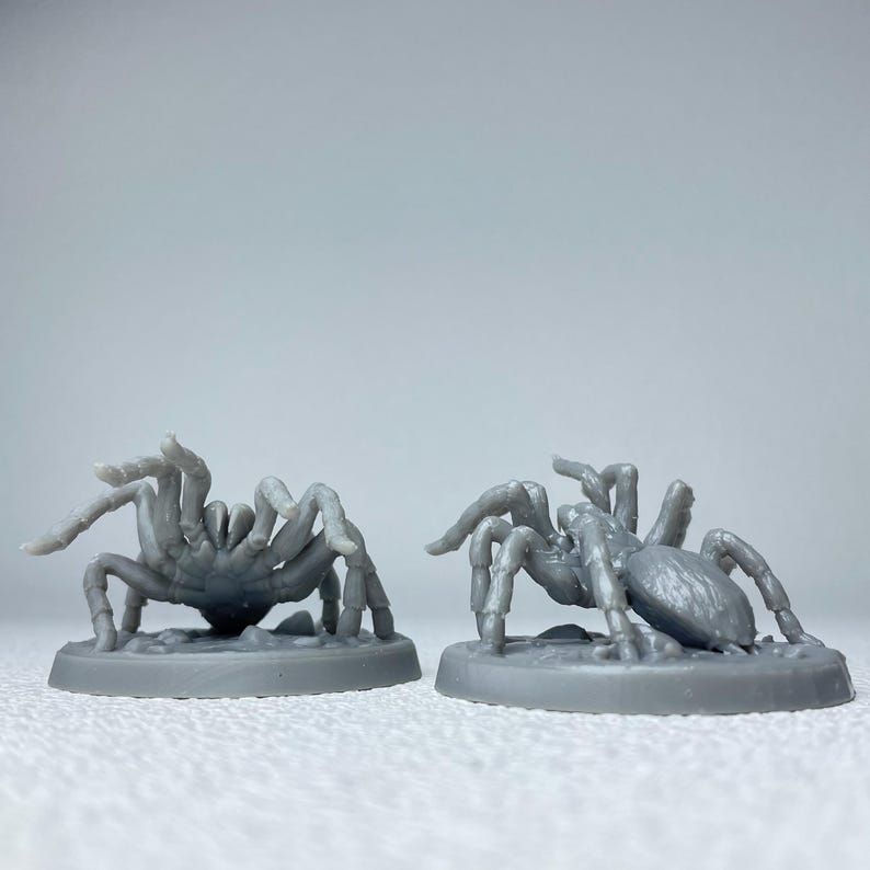 May include: Two gray miniature spider figurines. Each spider has eight legs and a detailed body. The spiders are on circular bases. The background is a solid, light gray color.
