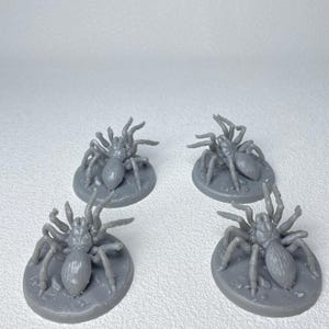 May include: Four gray miniature spider figurines, each with eight legs and a rounded body, are displayed on circular bases. The figurines are designed for tabletop gaming or crafting projects.