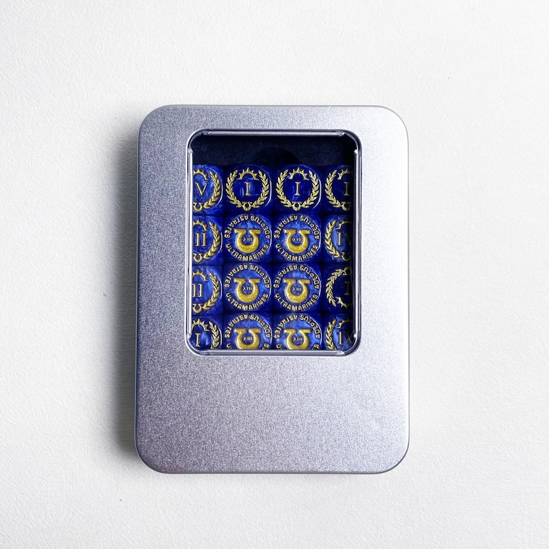 May include: A silver metal tin containing blue dice with gold Roman numerals and laurel wreaths. Each die features the text "ULTRAMARINES" and a symbol. The dice are arranged neatly inside the tin.