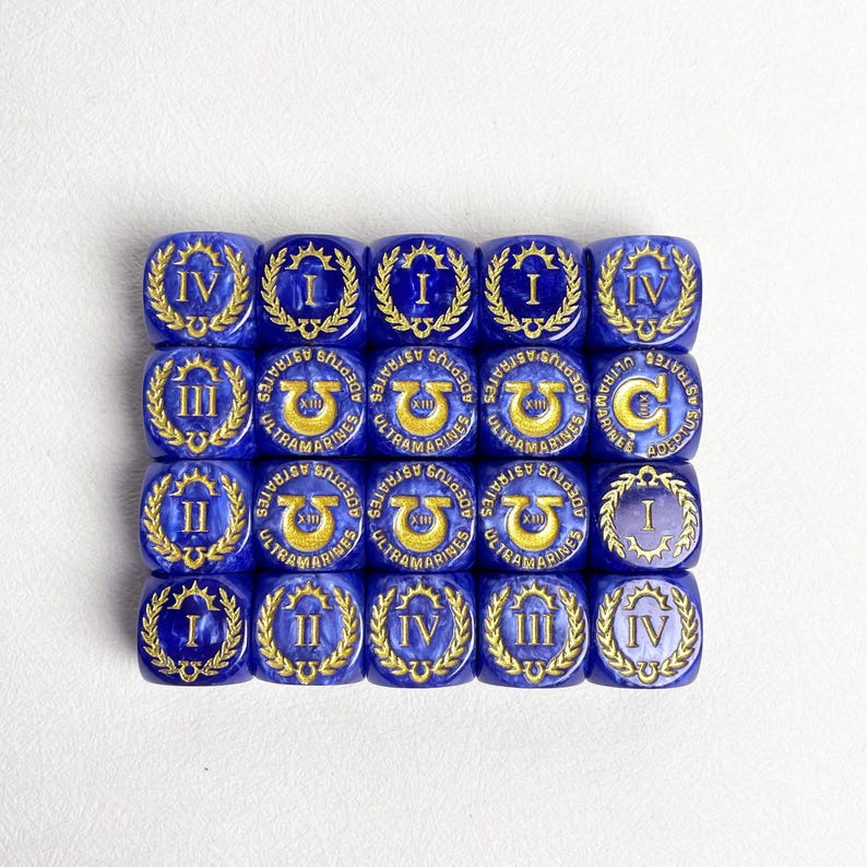 May include: A collection of twenty blue dice with gold Roman numerals and laurel wreaths. Each die features the text "ULTRAMARINES" and "ADEPTUS ASTRATES". The dice are arranged in a grid pattern on a white surface.