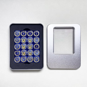 May include: A set of sixteen blue dice with gold markings, arranged in a black tray inside an open silver metal box. Each die features Roman numerals and a decorative emblem. The box is on a white surface.
