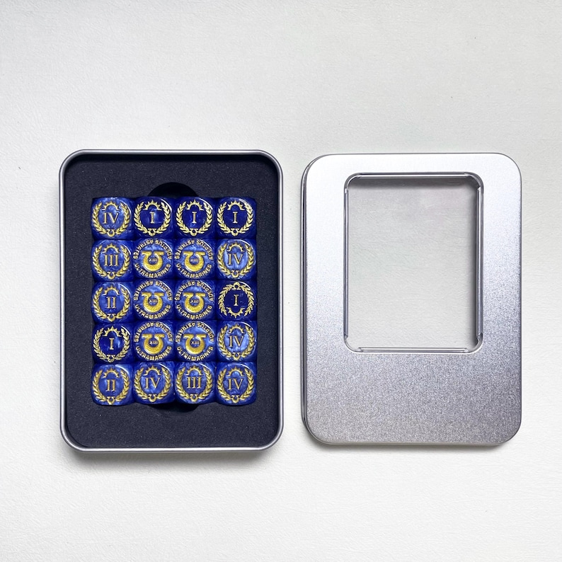 May include: A set of sixteen blue dice with gold markings, arranged in a black tray inside an open silver metal box. Each die features Roman numerals and a decorative emblem. The box is on a white surface.