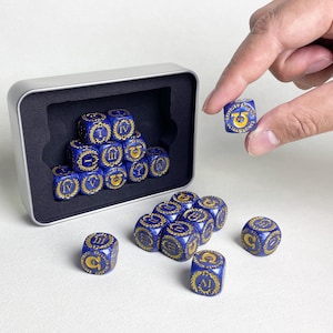 May include: A set of blue dice with gold detailing, featuring Roman numerals and symbols, presented in a silver tin. One die is held by a hand. The dice are for a tabletop game.