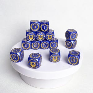 May include: A collection of blue dice with gold-colored Roman numerals and symbols. The dice are arranged on a white surface, with some stacked to form a small pyramid. The dice are a deep blue color with gold detailing.