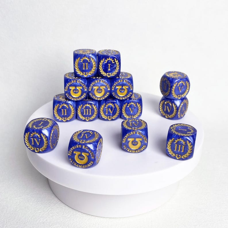 May include: A collection of blue dice with gold-colored Roman numerals and symbols. The dice are arranged on a white surface, with some stacked to form a small pyramid. The dice are a deep blue color with gold detailing.
