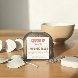 Peut inclure: Un pinceau lymphatique rectangulaire transparent avec le texte "Cowgirl Up" et "Lymphatic Brush" sur l'&eacute;tiquette. Le pinceau est &agrave; c&ocirc;t&eacute; d'un bol beige, d'un pain de savon et de brins de lavande sur une surface en bois.