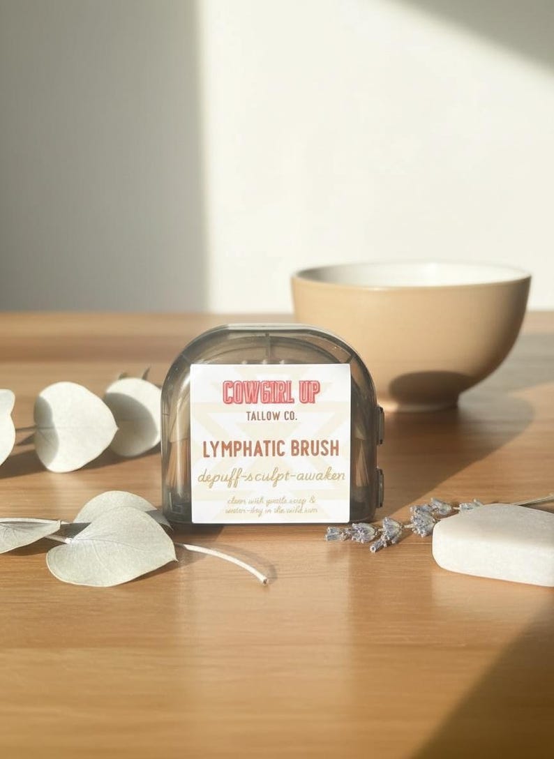 Peut inclure: Un pinceau lymphatique rectangulaire transparent avec le texte "Cowgirl Up" et "Lymphatic Brush" sur l'&eacute;tiquette. Le pinceau est &agrave; c&ocirc;t&eacute; d'un bol beige, d'un pain de savon et de brins de lavande sur une surface en bois.