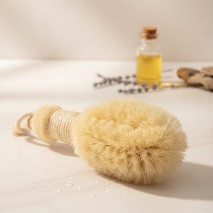Op de afbeelding: Een droge borstel met natuurlijke borstelharen en een houten handvat omwikkeld met touw. De borstel heeft een lus om op te hangen. Een klein flesje olie en lavendel takjes staan op de achtergrond. De borstel is voor lichaamsexfoliatie.
