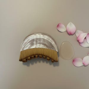 Peut inclure: Un pinceau de maquillage avec un manche incurv&eacute; et marbr&eacute; et un &eacute;ventail de poils dor&eacute;s. Un petit gel transparent et plusieurs p&eacute;tales de fleurs roses et blancs sont &agrave; c&ocirc;t&eacute; du pinceau sur une surface neutre.