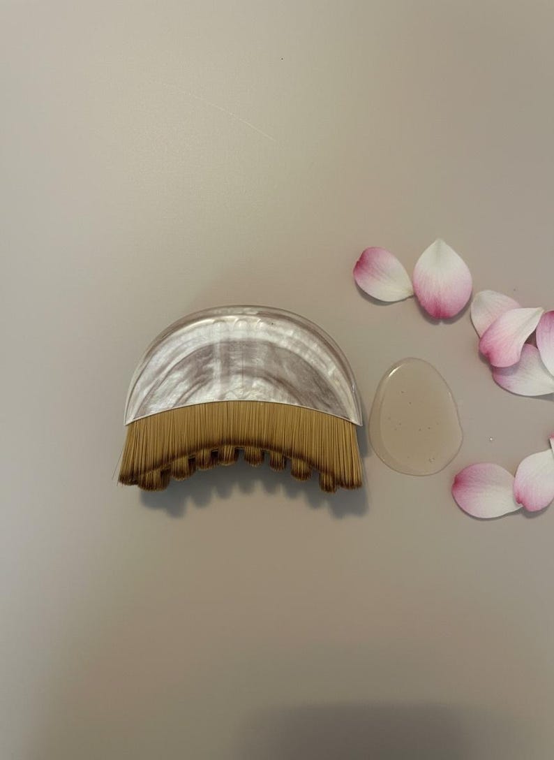 Peut inclure: Un pinceau de maquillage avec un manche incurv&eacute; et marbr&eacute; et un &eacute;ventail de poils dor&eacute;s. Un petit gel transparent et plusieurs p&eacute;tales de fleurs roses et blancs sont &agrave; c&ocirc;t&eacute; du pinceau sur une surface neutre.