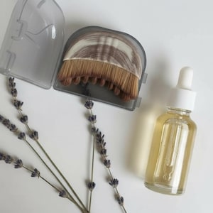 Peut inclure: Une bouteille en verre transparent avec un compte-gouttes blanc, remplie d'un liquide jaune, est pos&eacute;e &agrave; c&ocirc;t&eacute; d'un pinceau de maquillage dans un &eacute;tui gris. Des brins de lavande s&eacute;ch&eacute;e sont dispos&eacute;s sur une surface blanche.