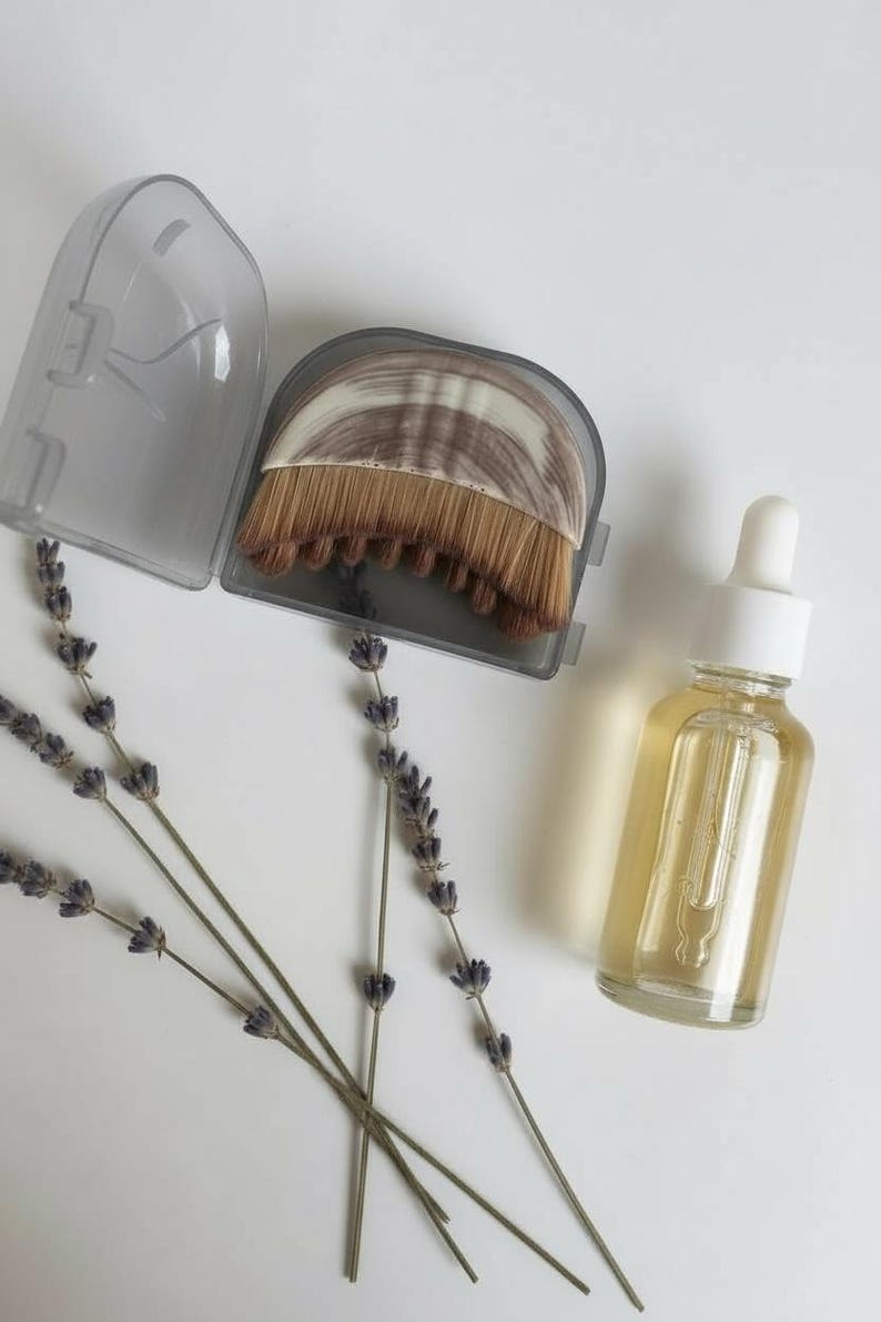 Peut inclure: Une bouteille en verre transparent avec un compte-gouttes blanc, remplie d'un liquide jaune, est pos&eacute;e &agrave; c&ocirc;t&eacute; d'un pinceau de maquillage dans un &eacute;tui gris. Des brins de lavande s&eacute;ch&eacute;e sont dispos&eacute;s sur une surface blanche.