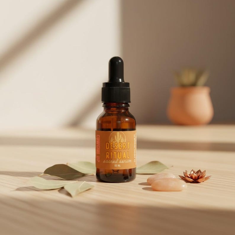 Peut inclure: Une bouteille en verre brun avec un bouchon compte-gouttes noir, &eacute;tiquet&eacute;e "Desert Ritual Sacred Serum". L'&eacute;tiquette pr&eacute;sente un paysage d&eacute;sertique. La bouteille est entour&eacute;e de feuilles, de pierres de quartz rose et d'une petite fleur cuivr&eacute;e sur une surface en bois.
