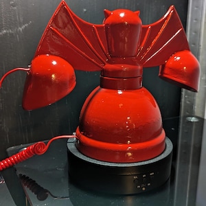 May include: A red novelty telephone with a bat-like design. The phone features a central body, two ear pieces, and wings. The phone sits on a black base with buttons and is connected by a red coiled cord.