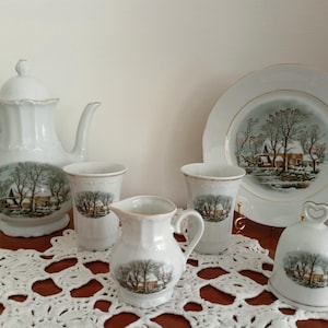 Puede incluir: Juego de té de porcelana blanca con un diseño de escena invernal. El juego incluye una tetera, tazas, una crema, un plato y una campana. La escena invernal presenta una casa, árboles y nieve.