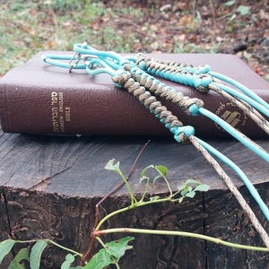 Puede incluir: Una Biblia de cuero marrón con texto en hebreo e inglés, sobre un tocón. Tres cordones trenzados en tonos azul, beige y marrón están sobre el libro. Follaje verde en primer plano, con un fondo borroso al aire libre.