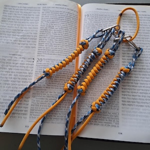Peut inclure: Ensemble de marque-pages faits à la main en paracorde jaune, bleu et blanc, avec des fermoirs en métal. Les marque-pages sont posés sur un livre ouvert avec du texte hébreu. Les marque-pages sont noués et tressés.