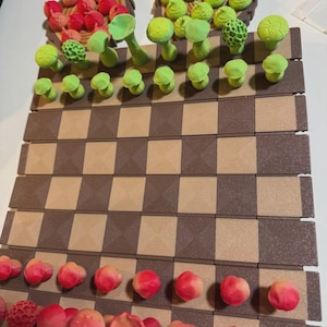 May include: A handmade chess set with a brown and tan checkered board. The chess pieces are whimsical, featuring red and pink berry-like shapes and green mushroom-shaped pieces. The pieces are arranged on the board, ready for play.