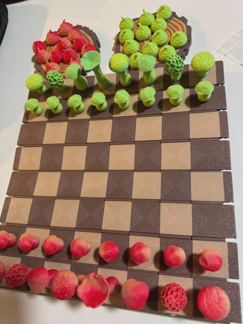 May include: A handmade chess set with a brown and tan checkered board. The chess pieces are whimsical, featuring red and pink berry-like shapes and green mushroom-shaped pieces. The pieces are arranged on the board, ready for play.