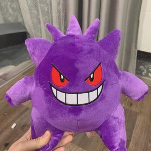 Pode incluir: Um brinquedo de pelúcia Gengar roxo com olhos vermelhos e um sorriso largo e ameaçador. O bicho de pelúcia tem saliências pontiagudas na cabeça e nos braços. Este peluche com tema Pokémon é um item colecionável popular.