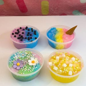 Pode incluir: Quatro recipientes de plástico transparente cheios de slime texturizado colorido. Um tem slime azul e roxo com contas pretas, outro tem slime azul, amarelo e rosa com um pequeno cone, um tem slime verde e rosa com flores, e um tem slime amarelo com cubos brancos.