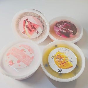 May include: Four round plastic containers filled with colourful slime, each featuring a circular label. The labels display the text Watermelon Drink, Dragon Fruit Cubes, Strawberry Milk, and Mango Boba, accompanied by illustrations of the respective fruits.