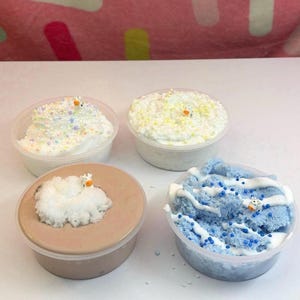 May include: Four clear plastic containers filled with textured slime. One contains white slime with pastel sprinkles, one white slime with yellow beads, one brown slime topped with white foam, and one blue slime with white swirls and blue beads.