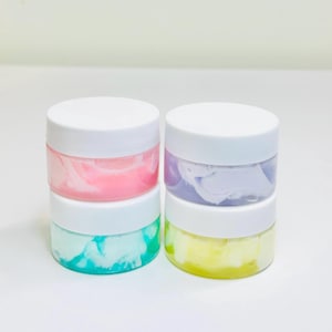 May include: Four small, clear jars with white lids. Each jar contains a different colored cream: pink, green, purple, and yellow. The creams have a marbled appearance. The jars are stacked in pairs.